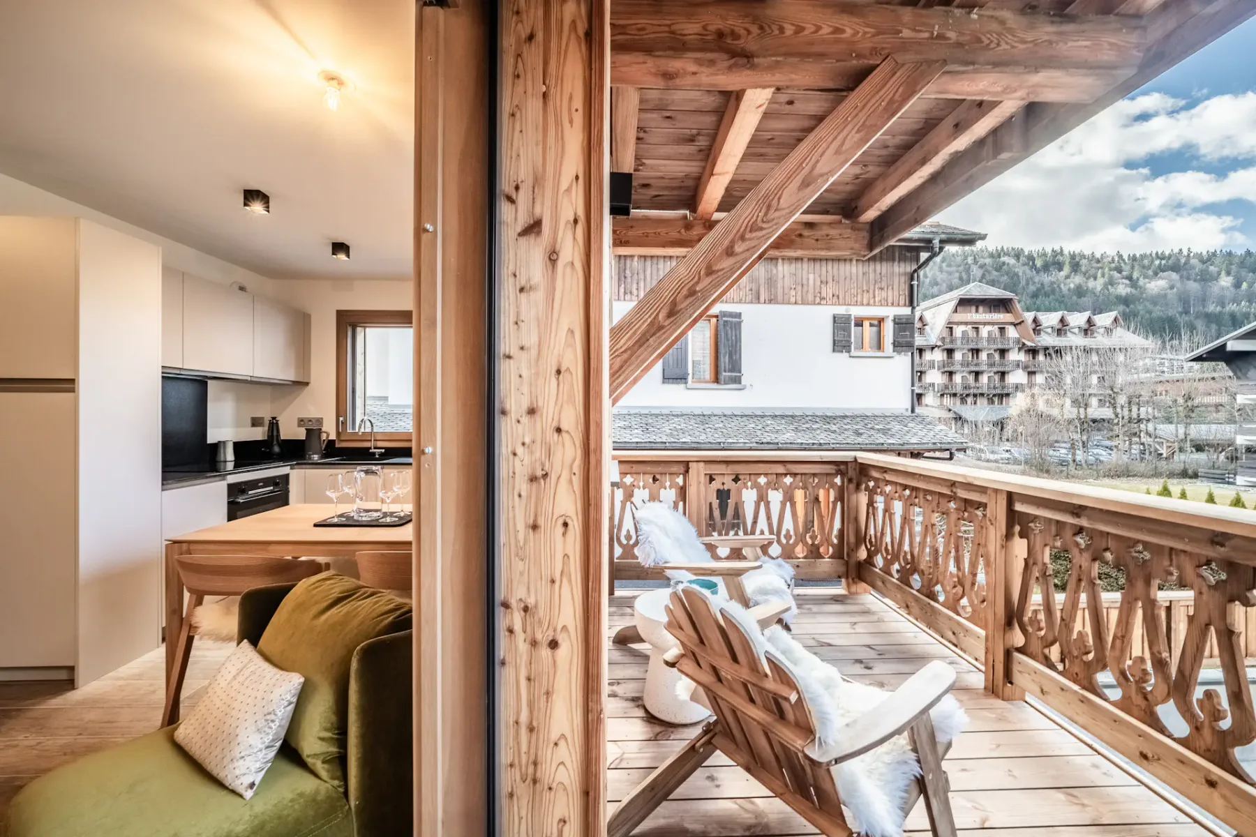 A rustic wooden balcony with lounge seating overlooks a village and wooded hills.