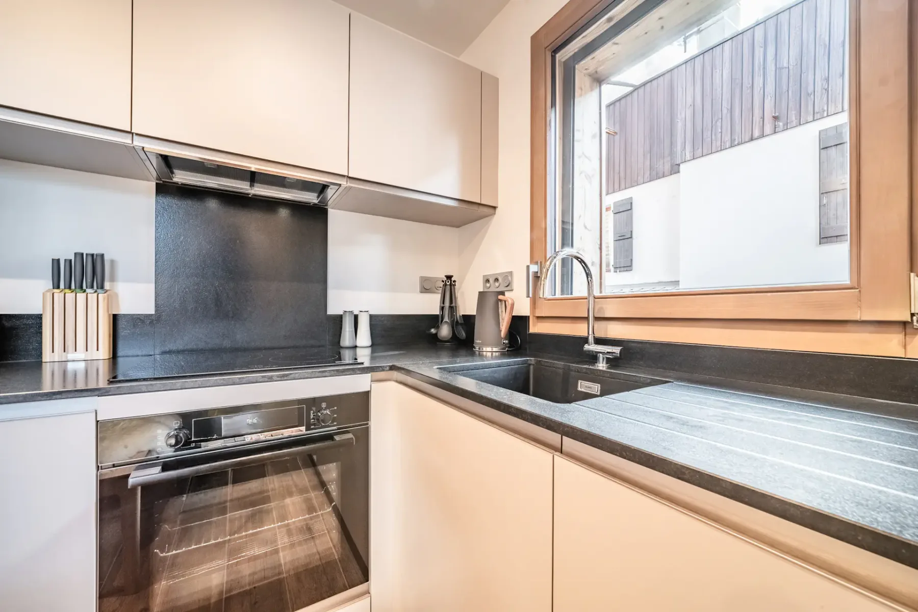 A modern kitchen with light cabinetry, dark countertops, and a sink by the window.