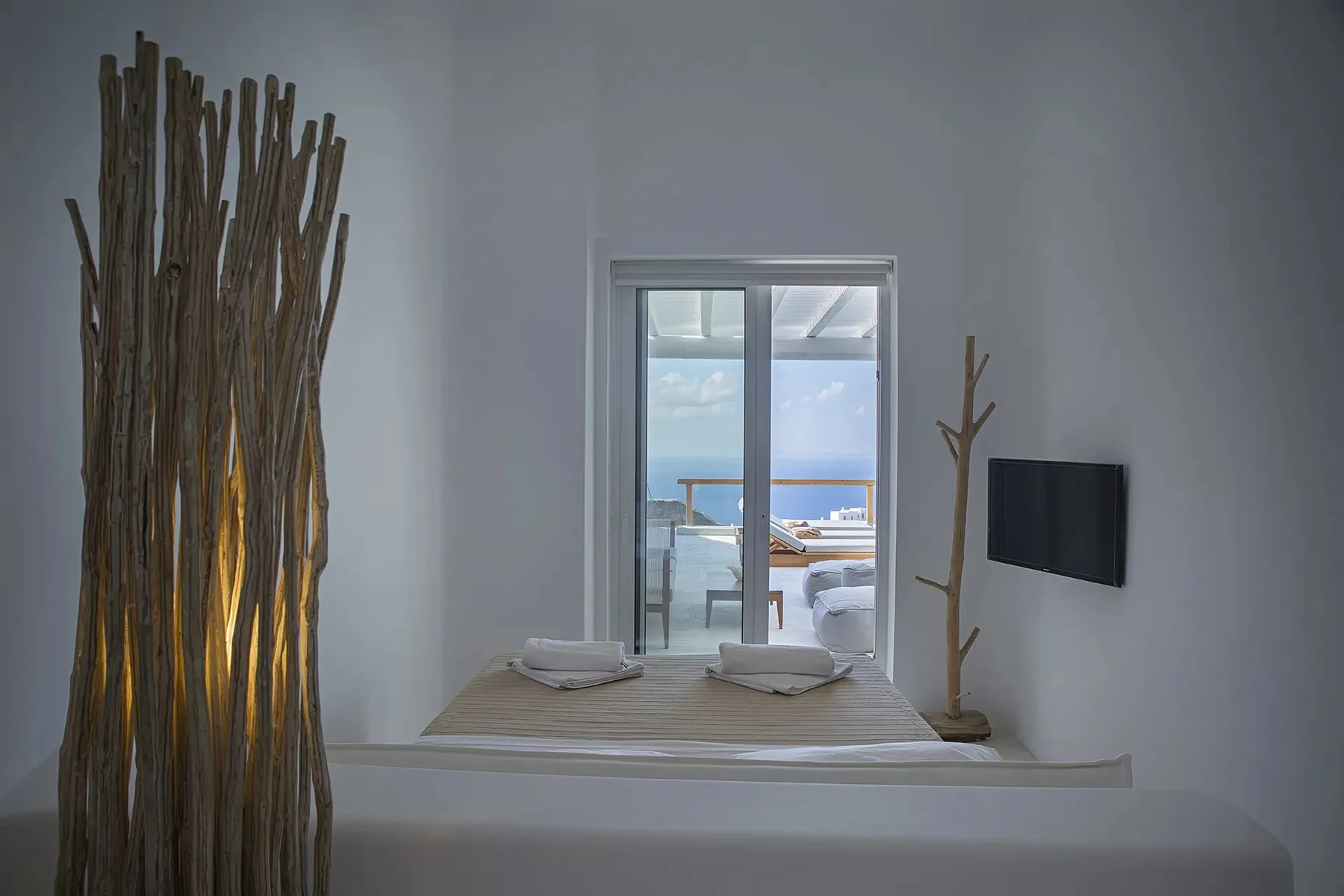 A bedroom with a neatly made bed, a wall-mounted television, and a view of the sea.