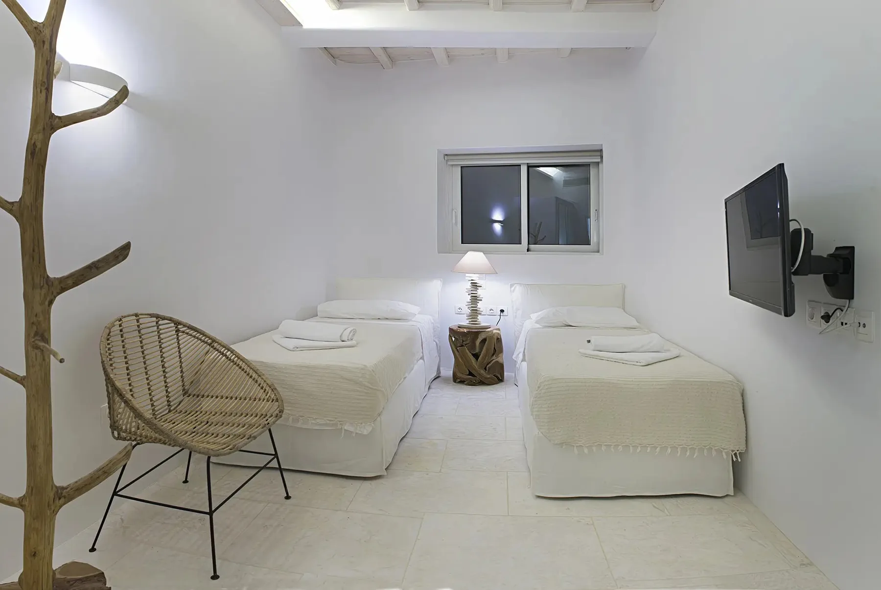 A bright bedroom with two single beds, a woven chair, and a wall-mounted television.
