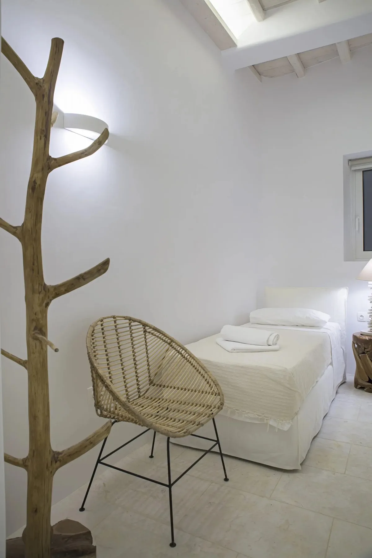 A bright bedroom with a bed and a woven chair beside a natural wood coat stand.
