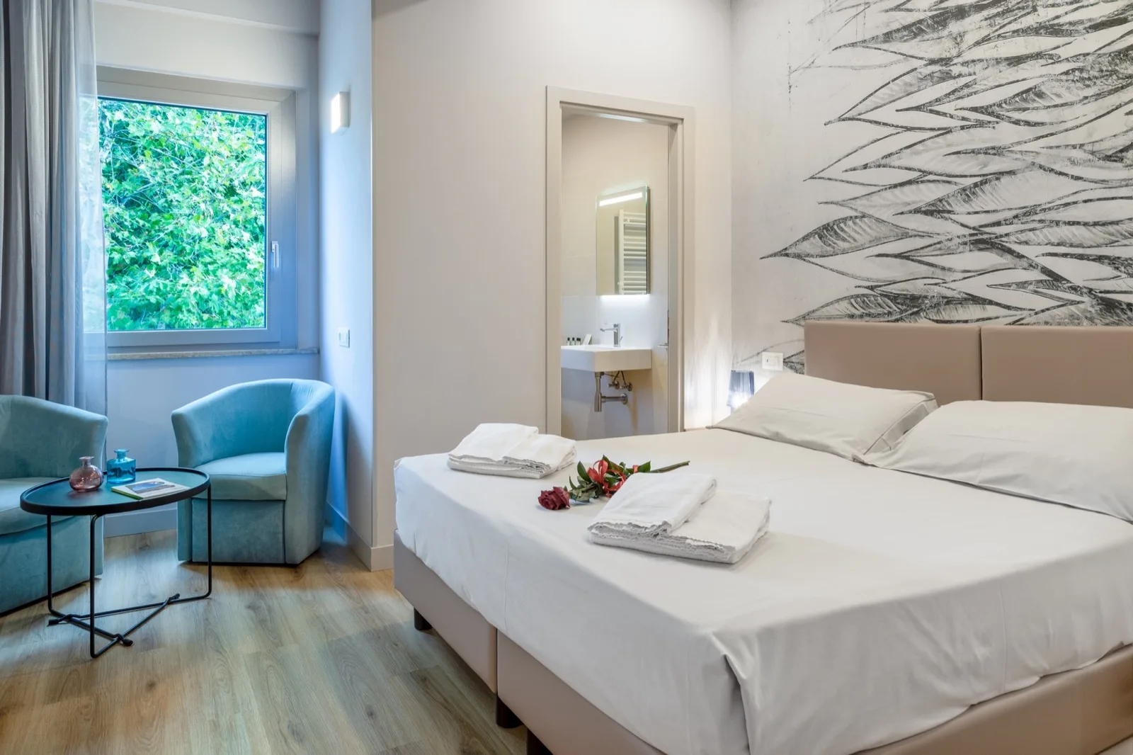 Representative hotel space: A bedroom with a large bed, a blue armchair, and a window overlooking greenery.