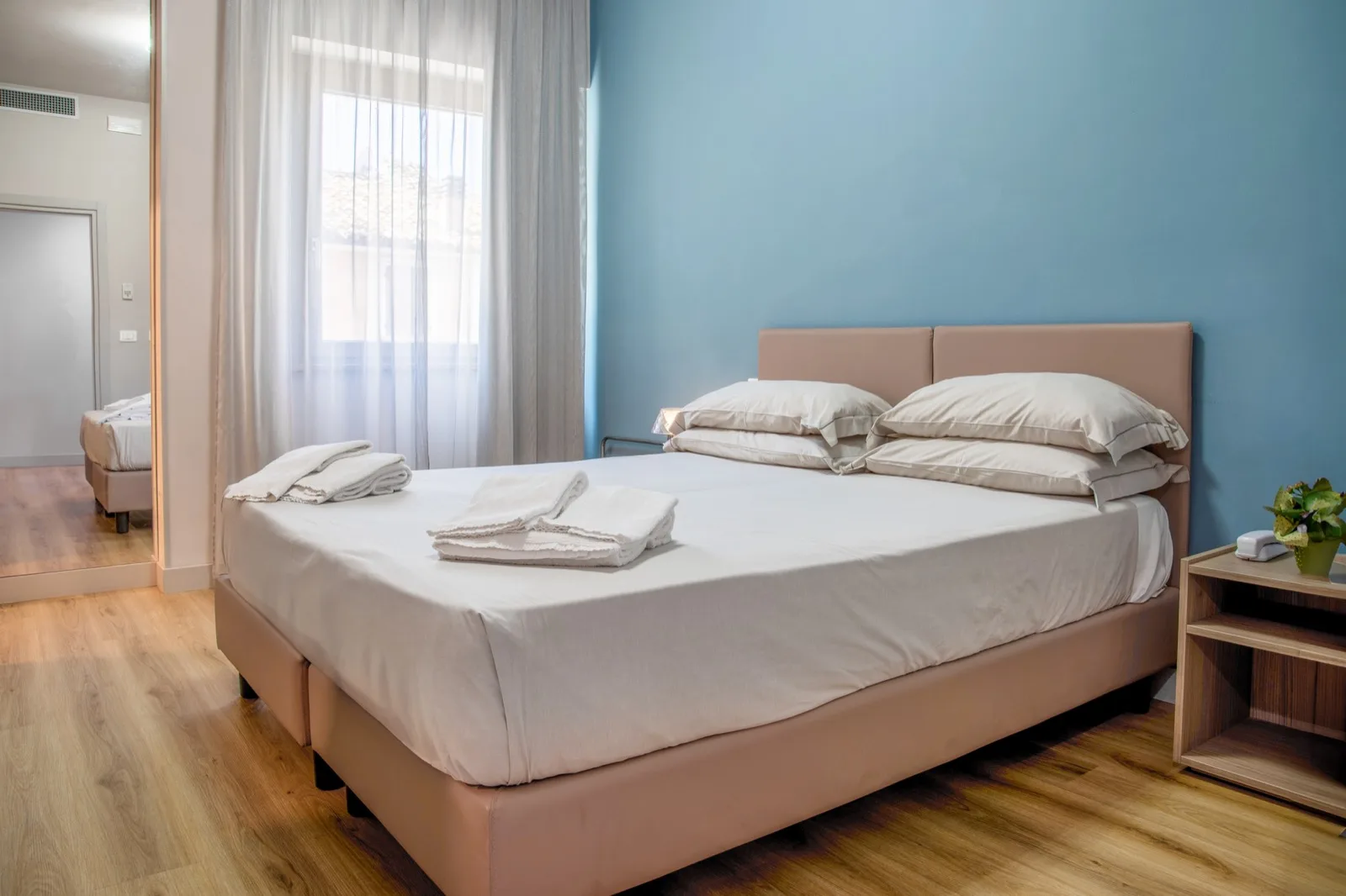 Representative hotel space: A bedroom with a neatly made bed beside a window with sheer curtains.