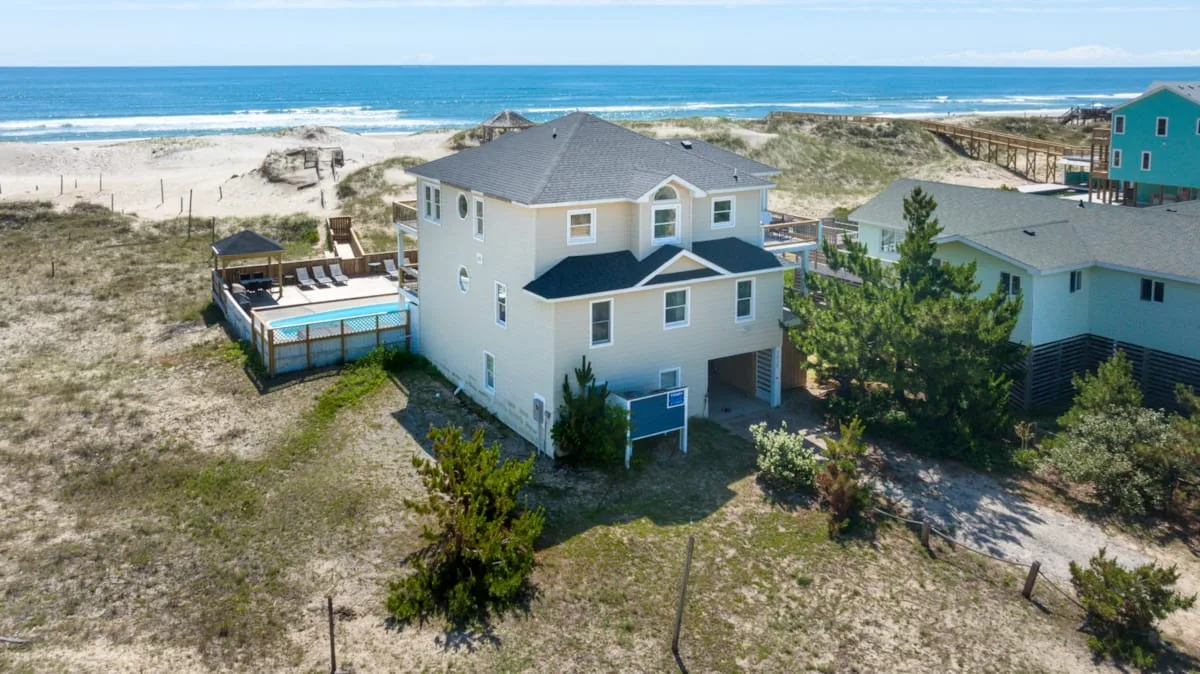 Aerial view of a coastal house surrounded by sandy dunes, with the ocean beyond.