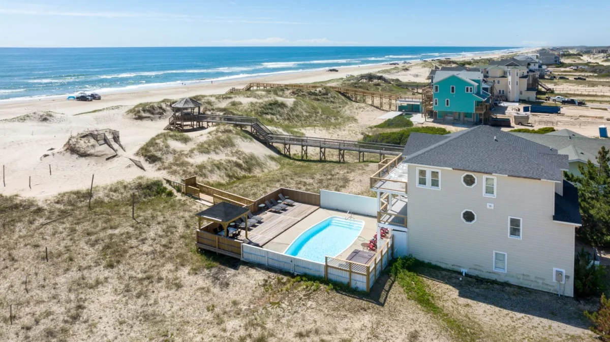A coastal house with a pool sits beside sandy dunes and the ocean.