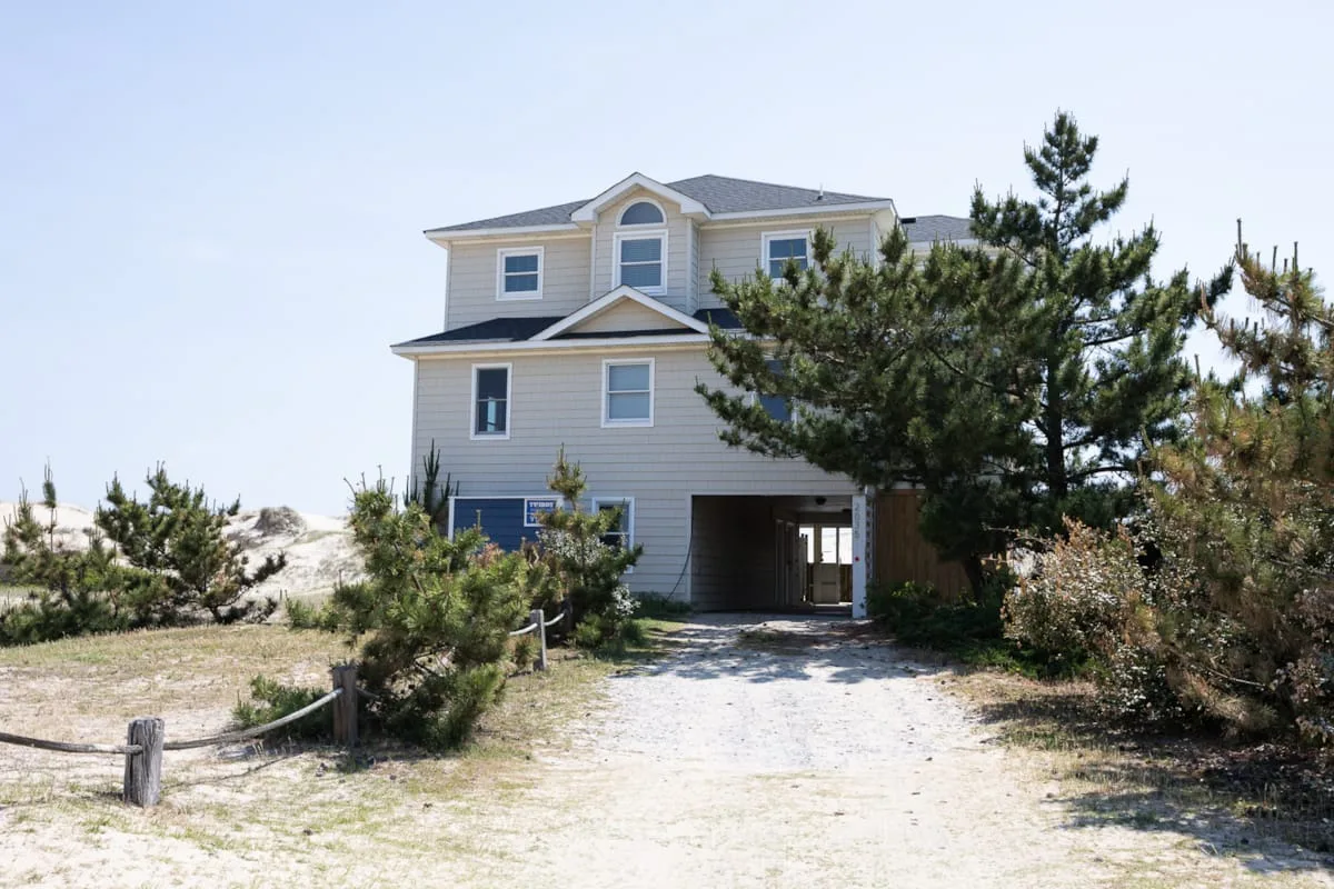 A large light-colored house stands at the end of a sandy driveway among trees.