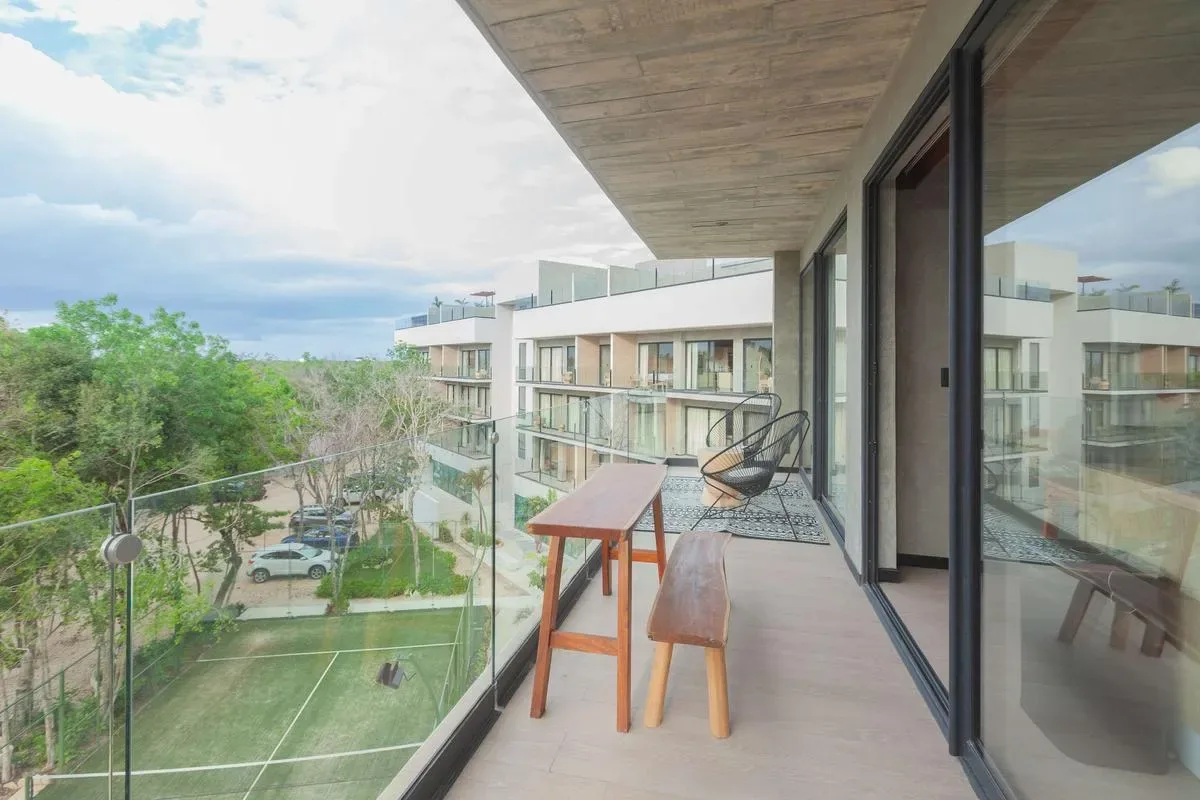 A covered balcony with a glass railing, wooden table and stools overlooks landscaped grounds and nearby buildings.