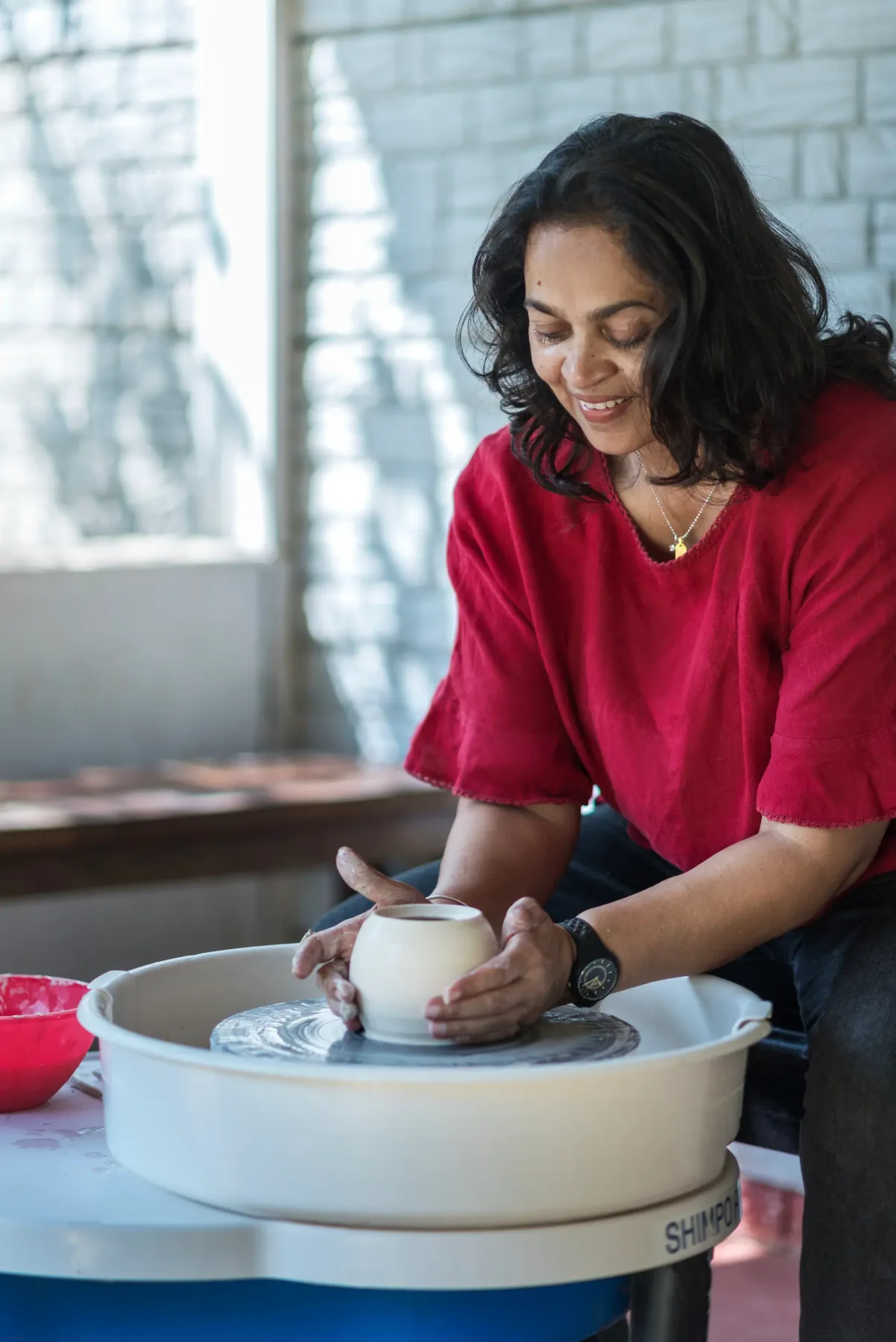 A woman shapes a ceramic vessel on a pottery wheel in a bright studio.