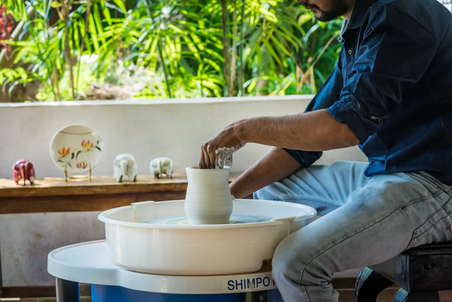 A person shapes a clay vessel on a pottery wheel in a garden-side workspace.