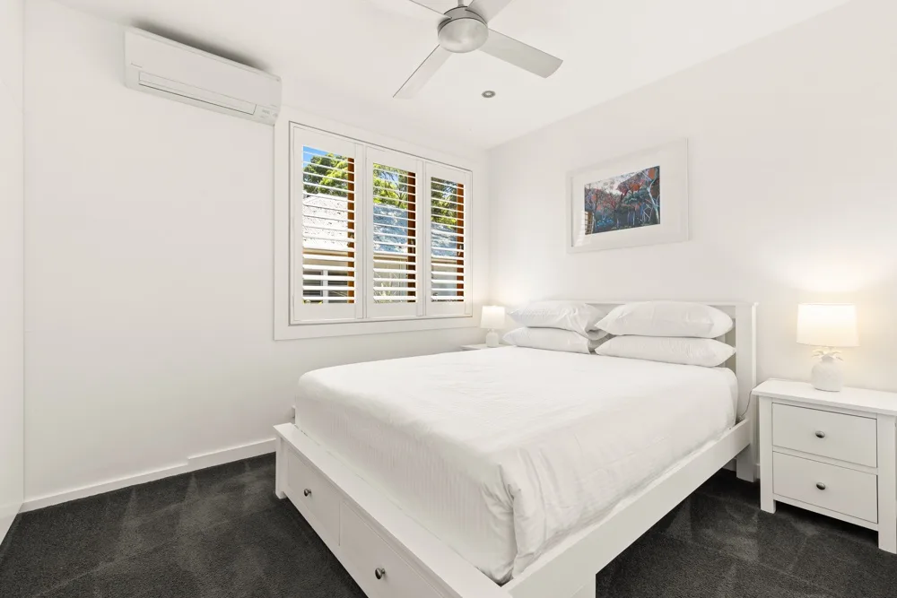 Un dormitorio luminoso con una cama blanca bajo una ventana con persianas.