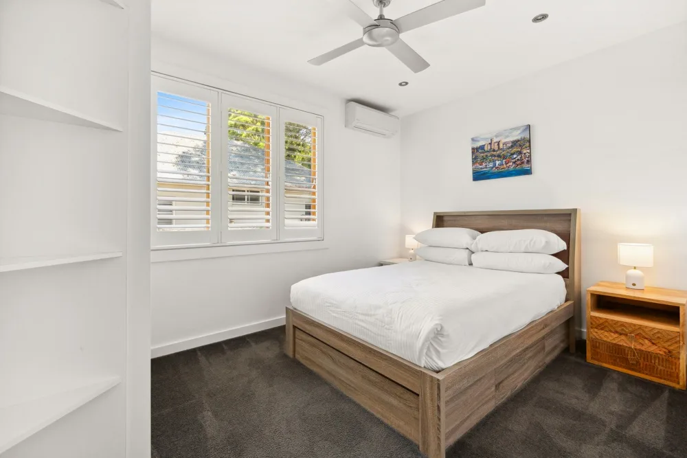 Un dormitorio luminoso con una cama de madera, ropa de cama blanca y ventanas con persianas.