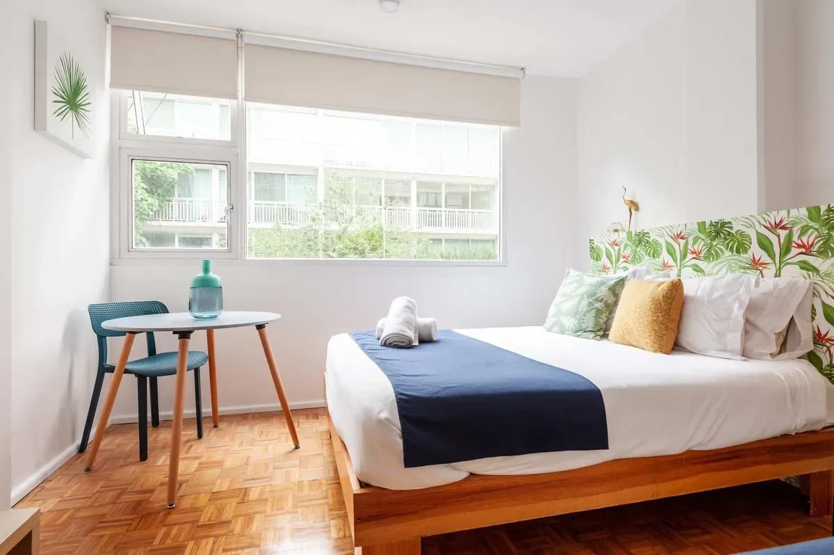 A bright bedroom features a wooden bed with blue bedding, leafy pillows, and a small table beside the windows.