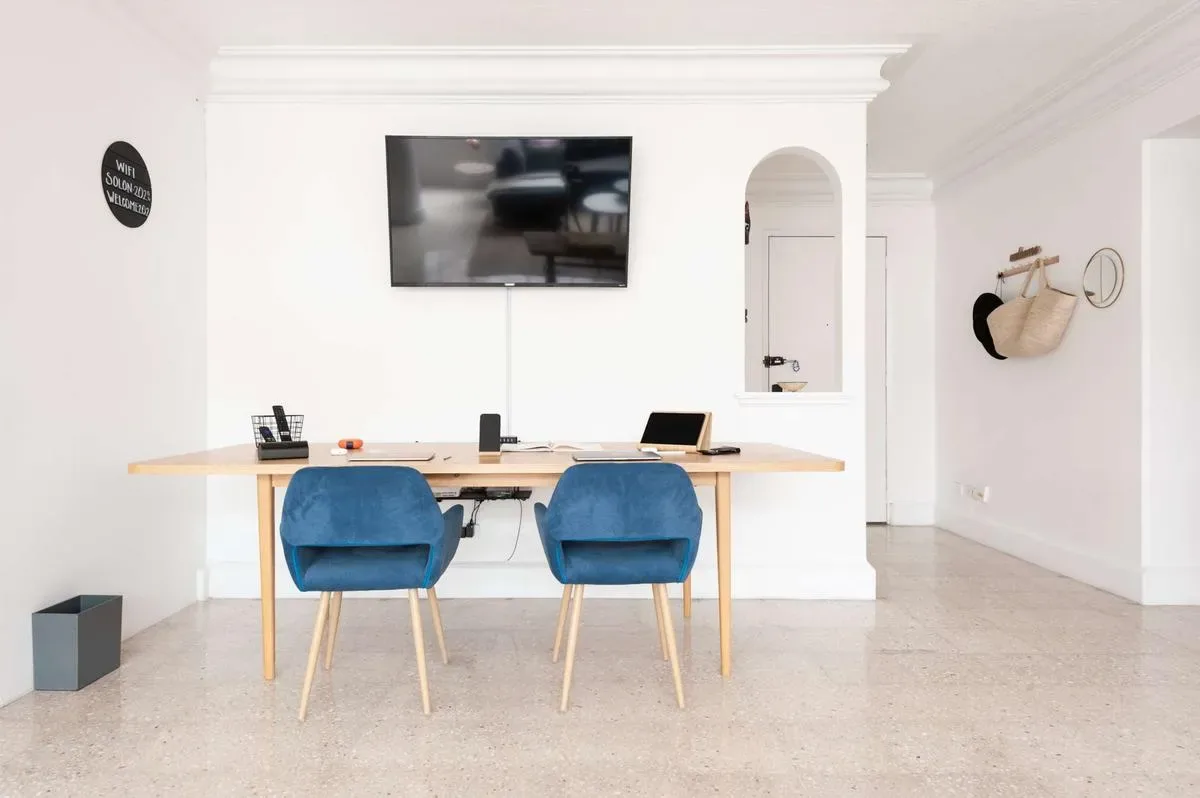 A bright white room features a long wooden desk, two blue chairs, and a wall-mounted television.