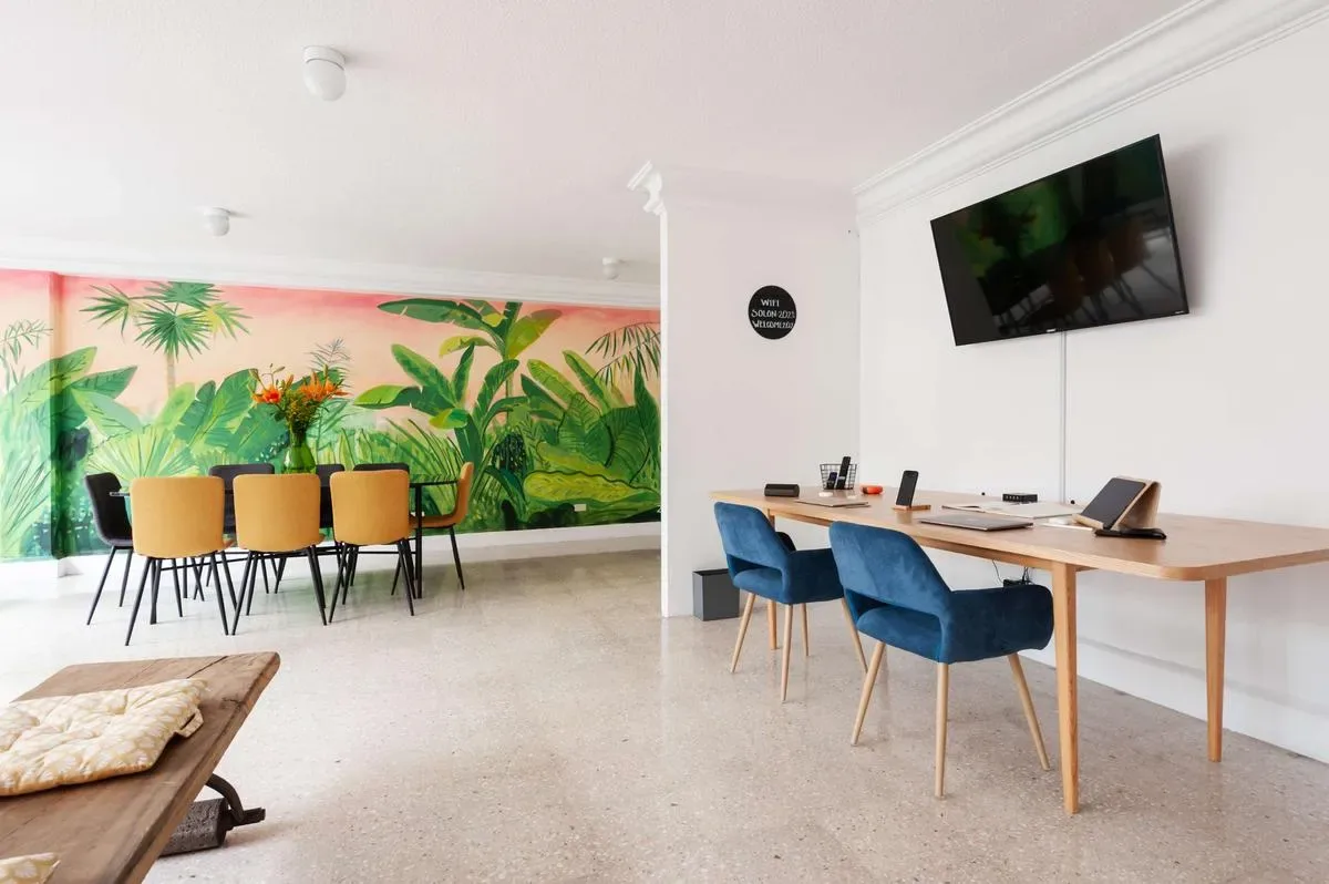 A bright lounge has a long wooden desk with blue chairs and a dining table against a leafy mural.
