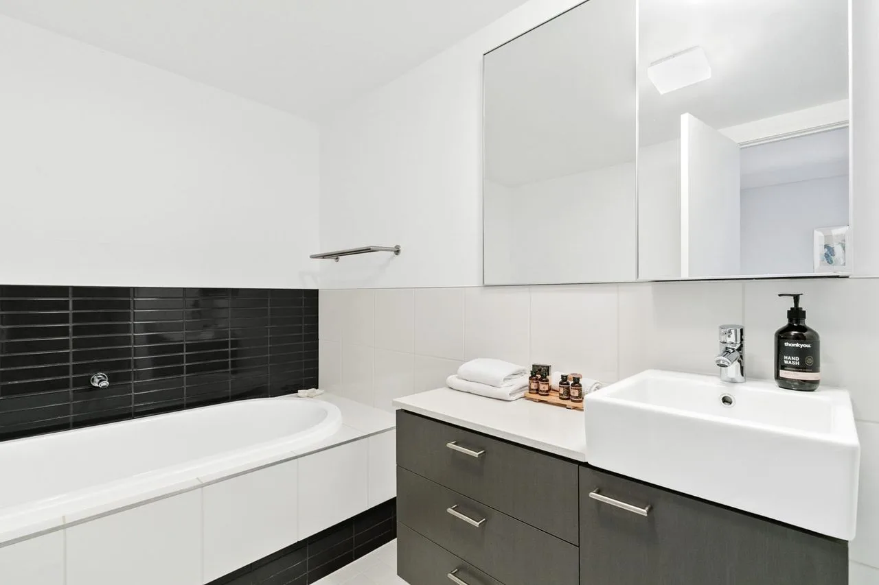 Baño luminoso con bañera, azulejos negros y un mueble con lavabo.