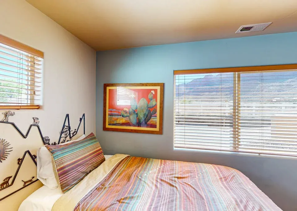 Un dormitorio con una cama colorida, una obra enmarcada y ventanas con persianas.