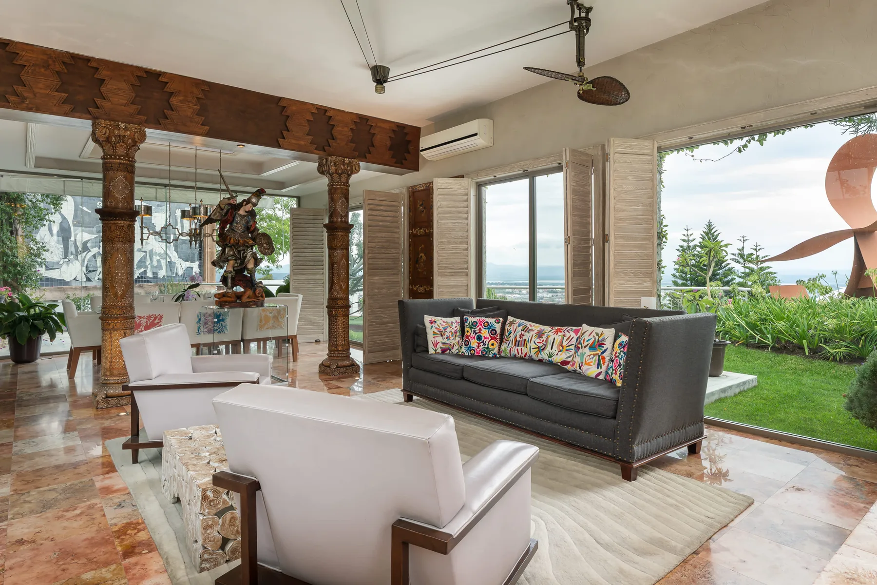 A bright living room with seating, a patterned sofa, and large windows overlooking greenery.
