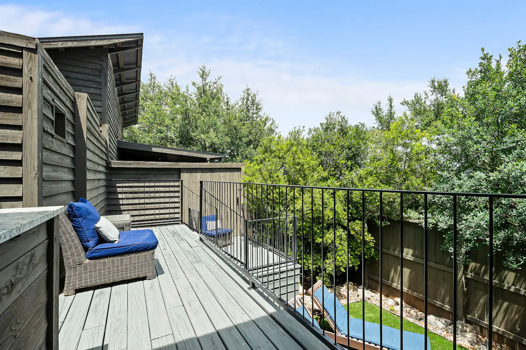 Una terraza de madera con asientos azules da a una zona exterior cercada y rodeada de árboles.