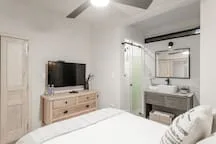 A bright bedroom has a bed, a dresser with a television, and an adjoining bathroom.