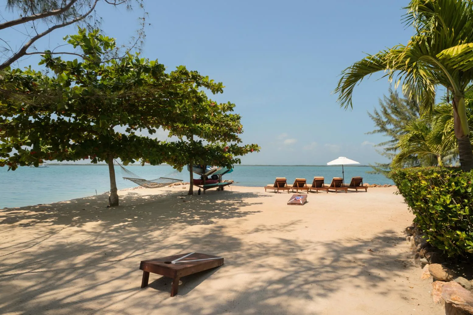 A sandy beach opens onto calm water, with shaded seating beneath trees.
