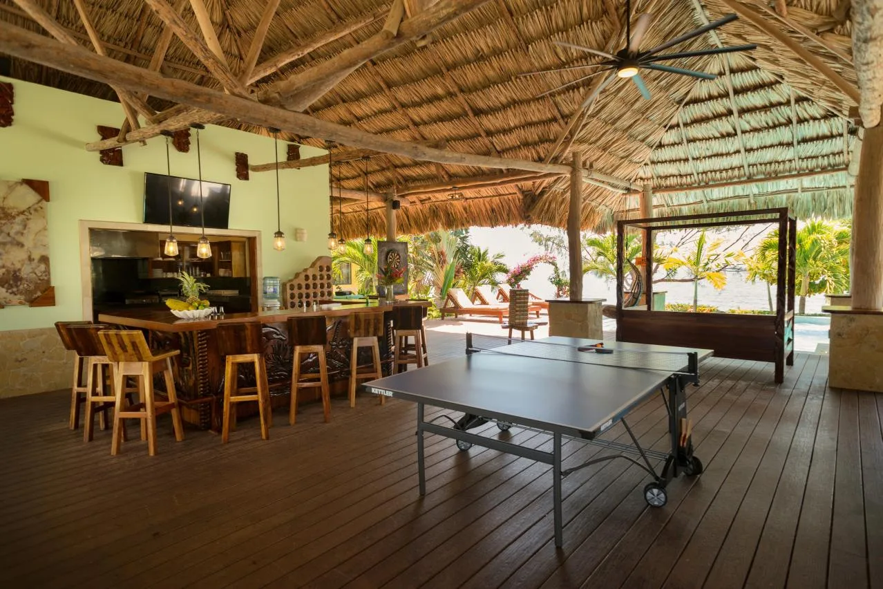 An open-air recreation area has a table tennis table, bar seating, and a thatched roof.