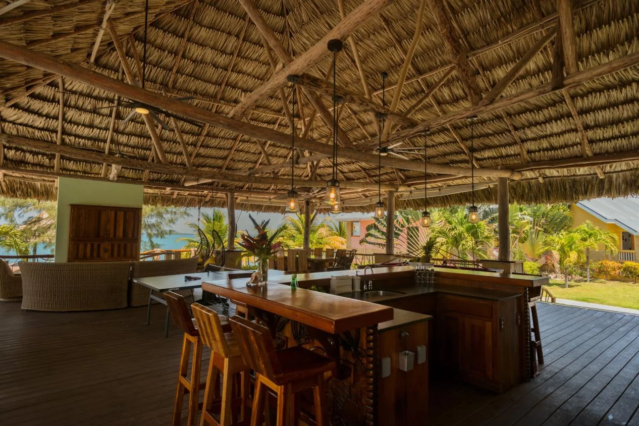 An open-air bar and dining area sits beneath a thatched roof, with greenery beyond.