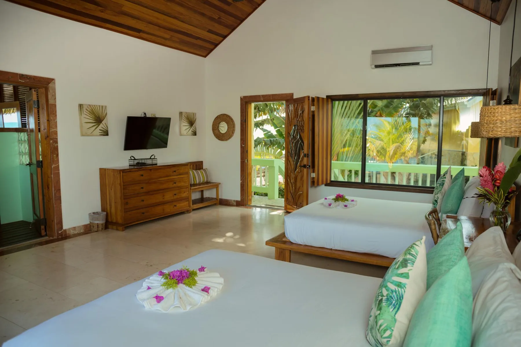 A bright bedroom with two beds and large windows overlooking tropical greenery.
