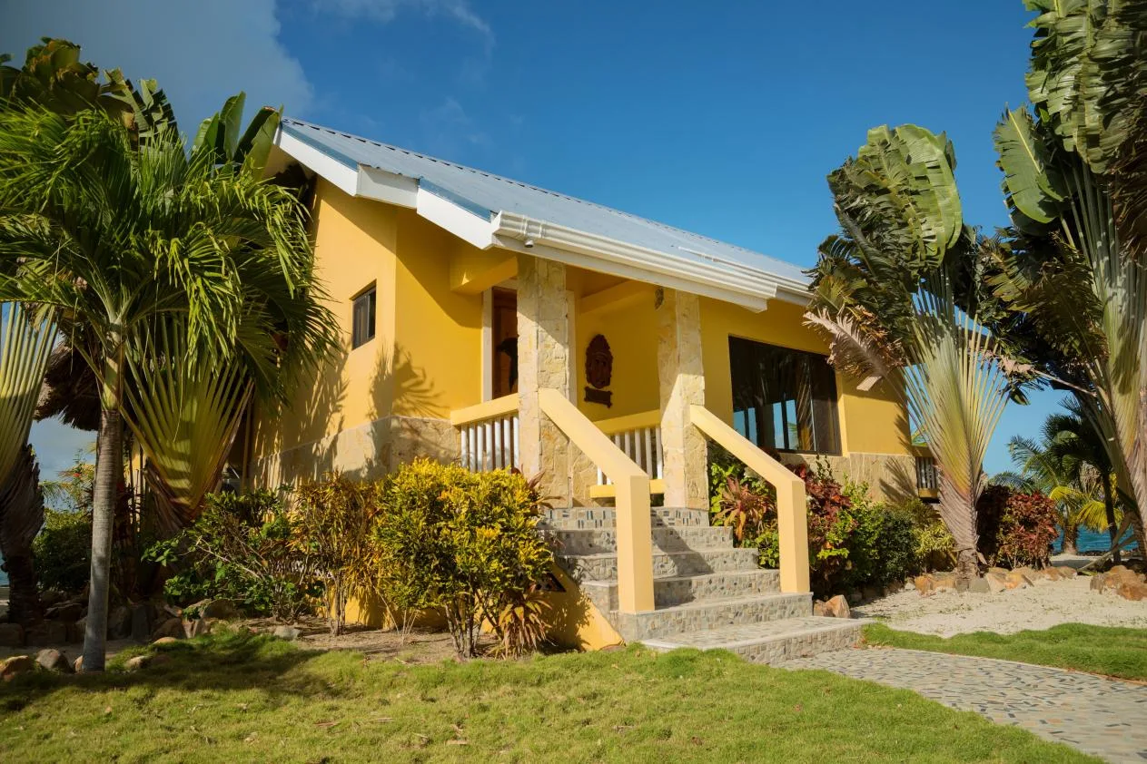 A yellow house stands among tropical plants, with steps leading to its entrance.