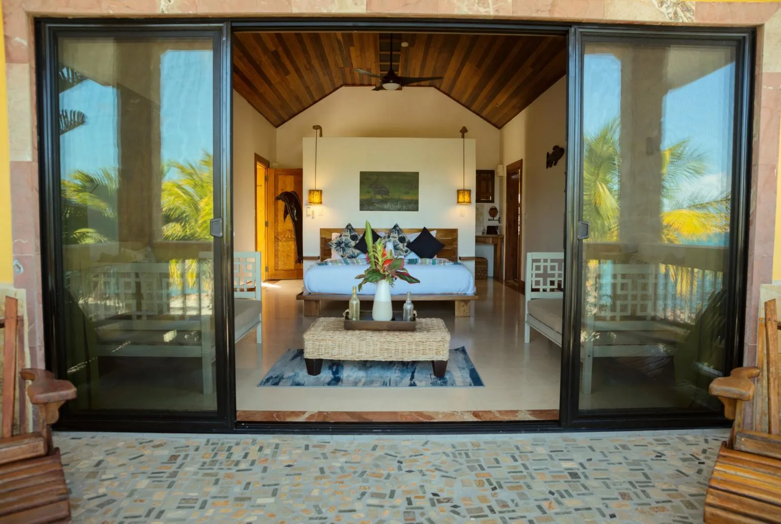 A bedroom opens onto a patio, with a bed beneath a high wooden ceiling.