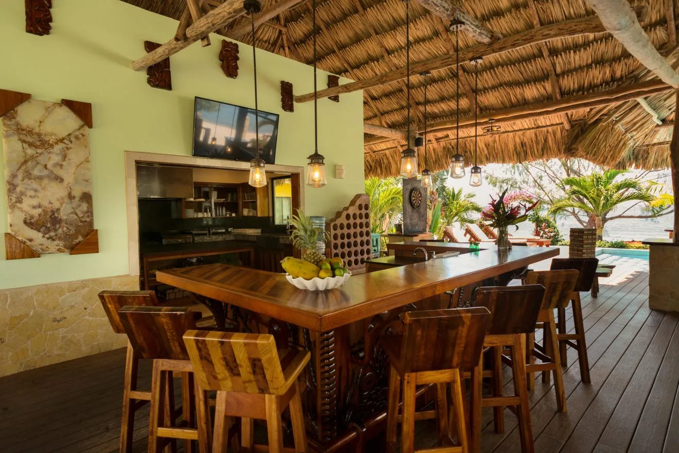 An open-air dining area with a long wooden table beneath a thatched roof.