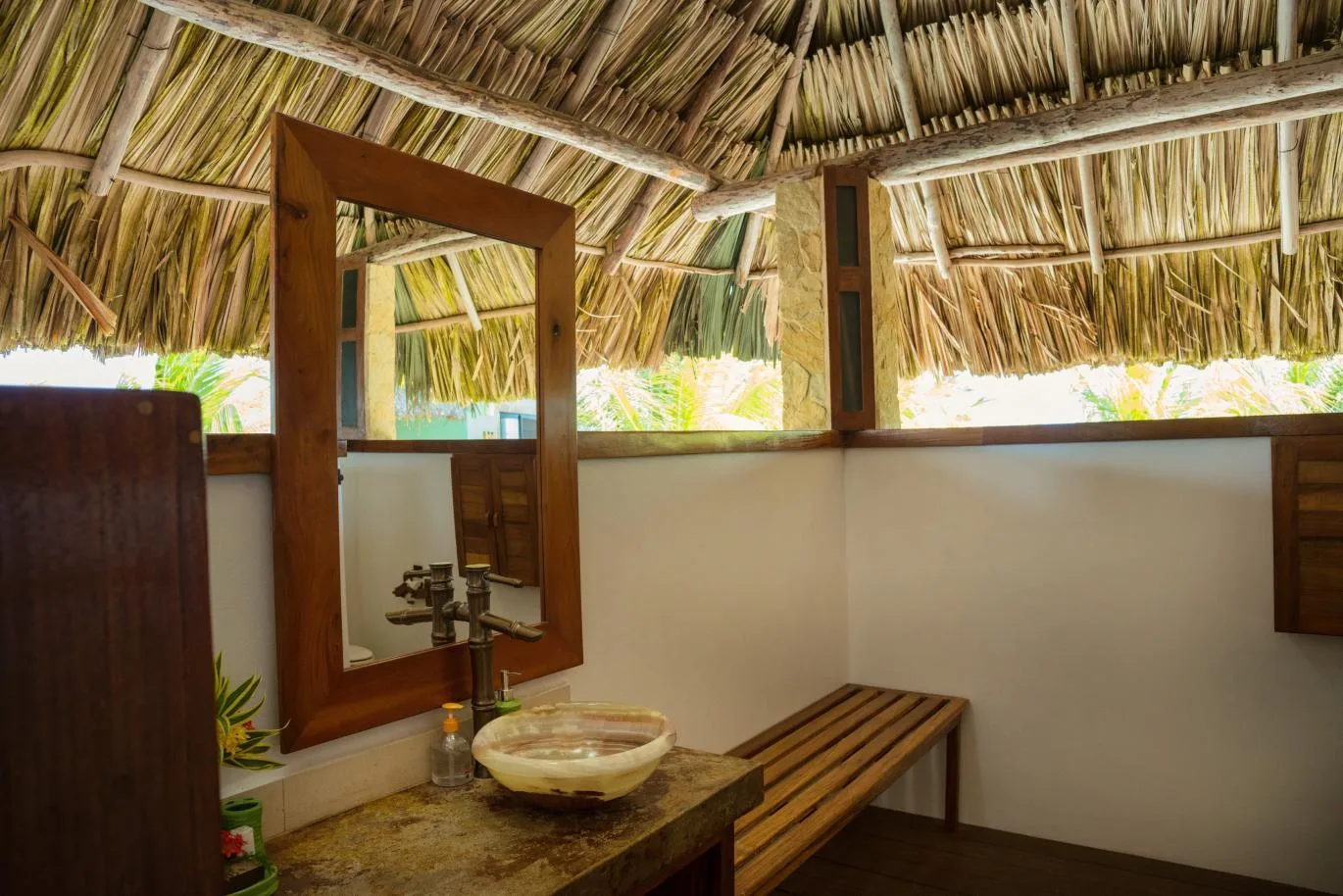 An open-sided bathroom with a thatched roof, a basin, and a wooden bench.