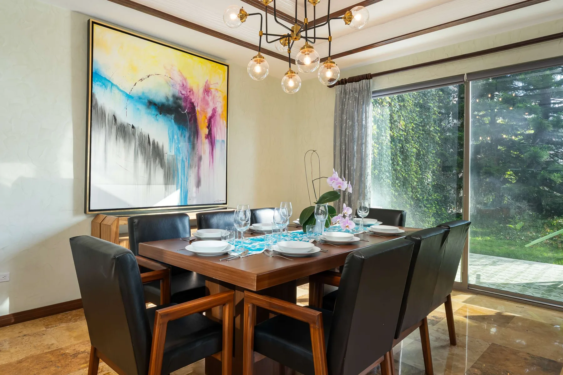 Comedor con la mesa puesta, asientos oscuros y una colorida obra de arte.