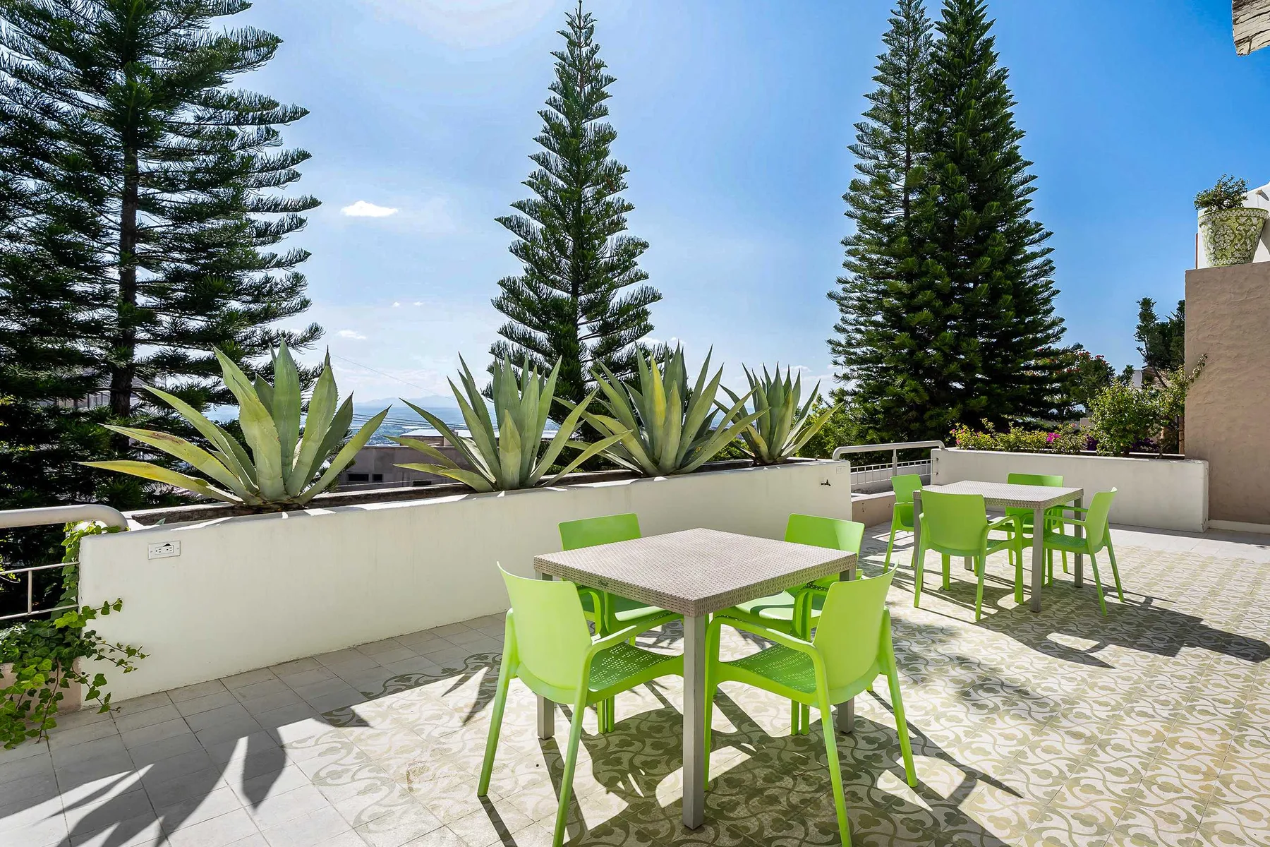 Un patio al aire libre con asientos verdes y árboles altos detrás de una jardinera baja.