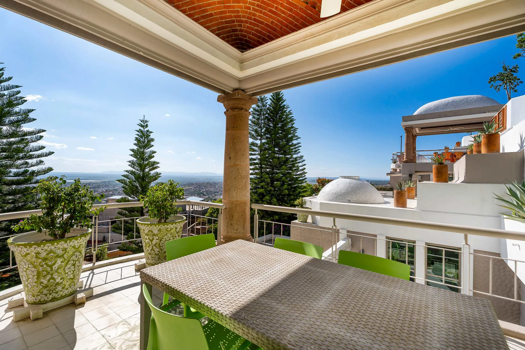 Una terraza luminosa al aire libre tiene una mesa, asientos verdes y vistas de árboles y tejados.