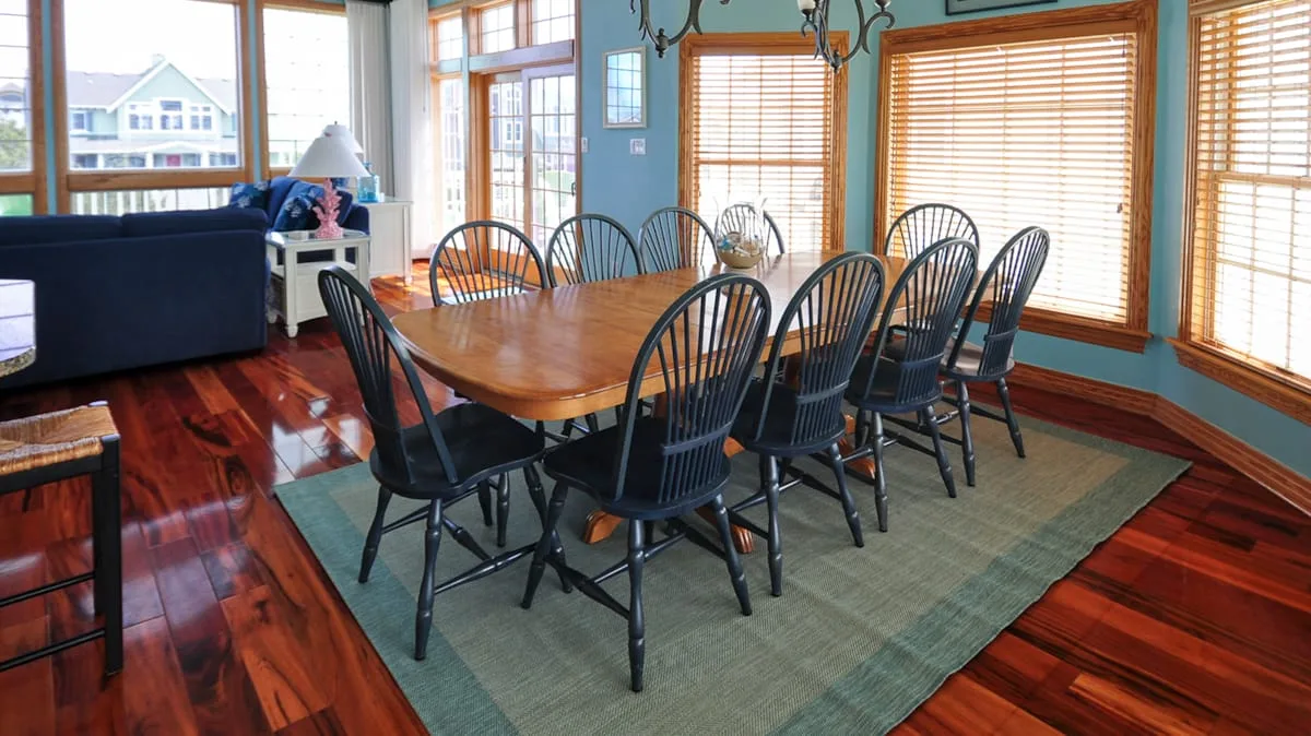 A dining area with a long wooden table and chairs beside large windows.