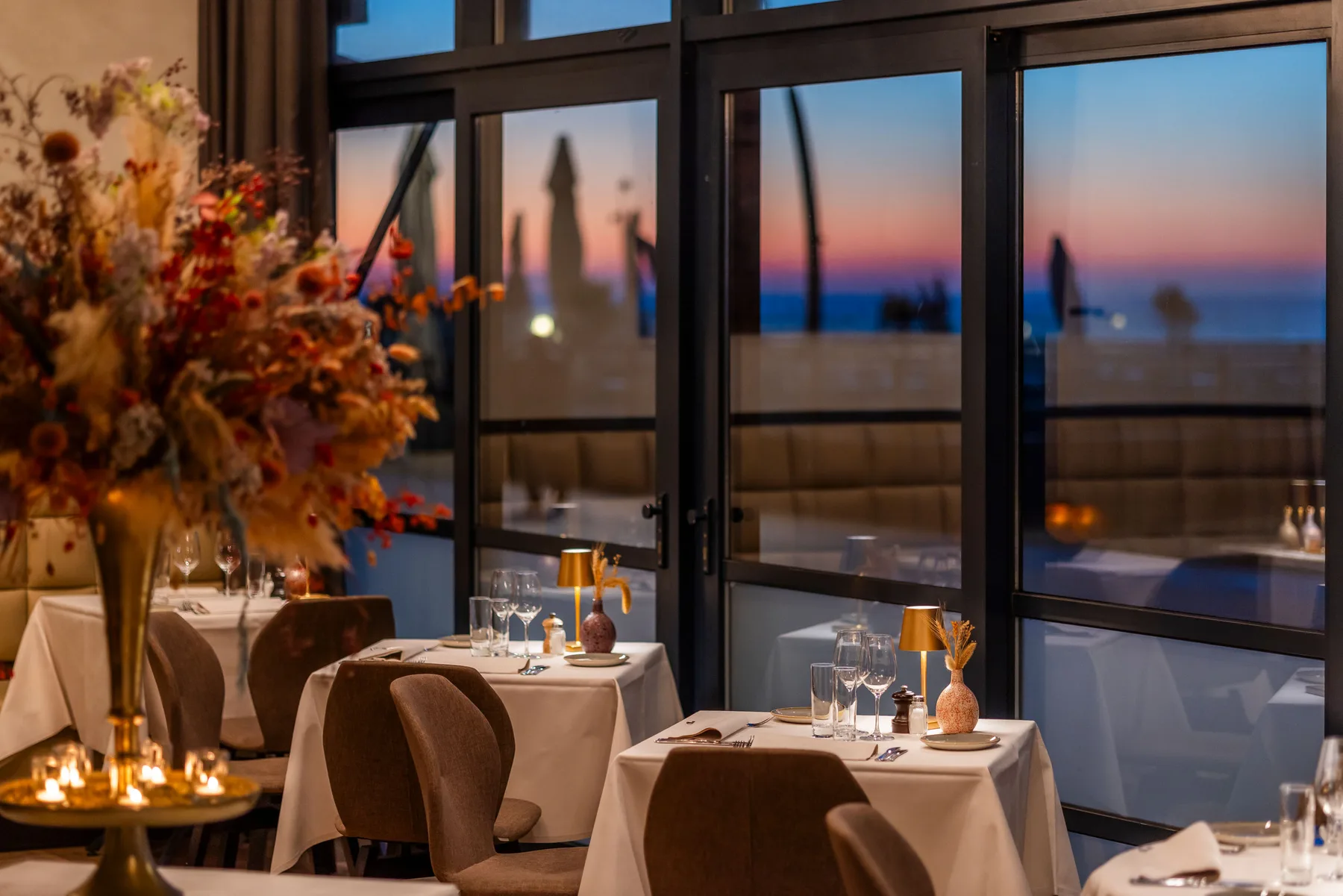 Espacio representativo del hotel: Un comedor con mesas preparadas junto a grandes ventanales que dan al agua al anochecer.