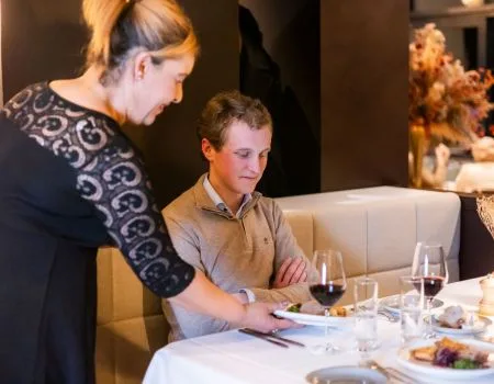 Espacio representativo del hotel: Una camarera atiende a un comensal sentado en una mesa servida con comida y vino.