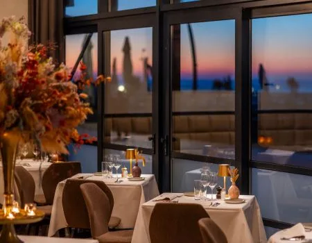 Espacio representativo del hotel: Comedor de restaurante con mesas preparadas junto a grandes ventanales al atardecer.