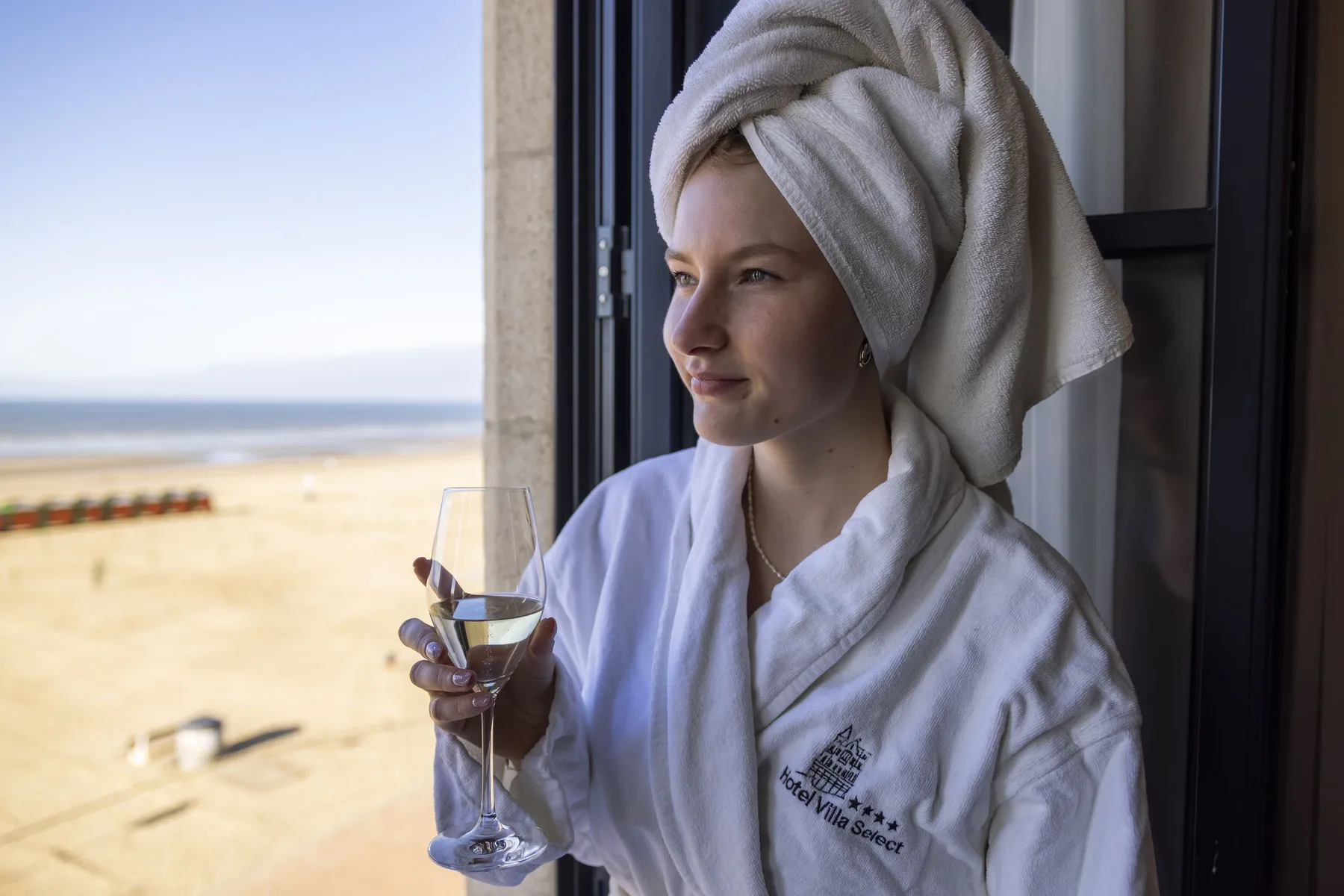 Espacio representativo del hotel: Una persona con bata y toalla enrollada sostiene una copa junto a una vista de playa.