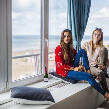 Espacio representativo del hotel: Dos mujeres están sentadas junto a una ventana grande con bebidas y vistas al mar.