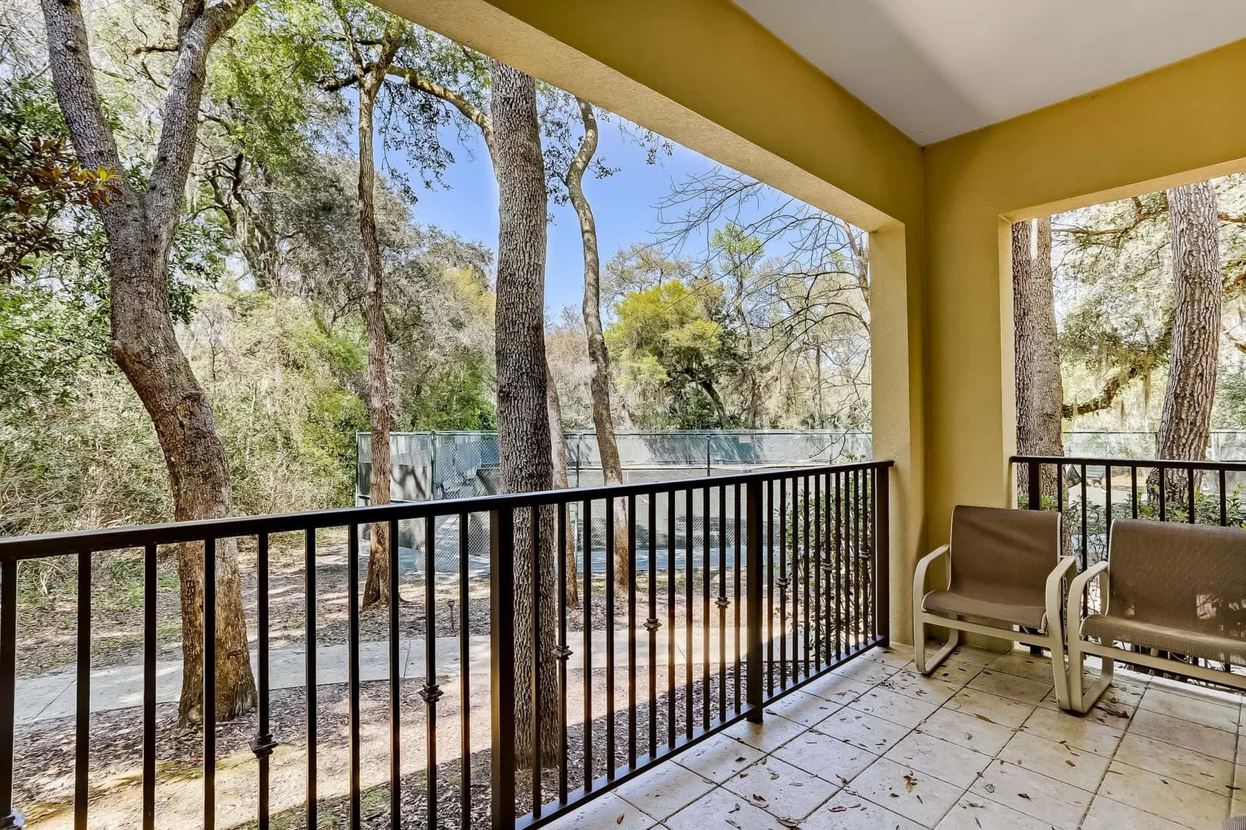 A covered outdoor balcony with metal railings and chairs overlooks a wooded area.