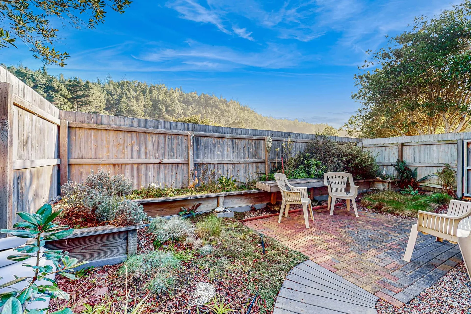 An outdoor patio has paved seating areas beside a wooden fence and landscaped garden.