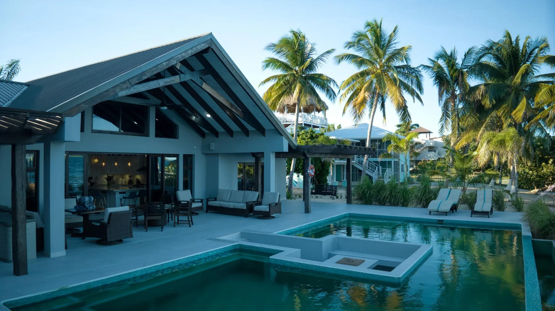 A modern villa opens onto a swimming pool, with palm trees in the background.