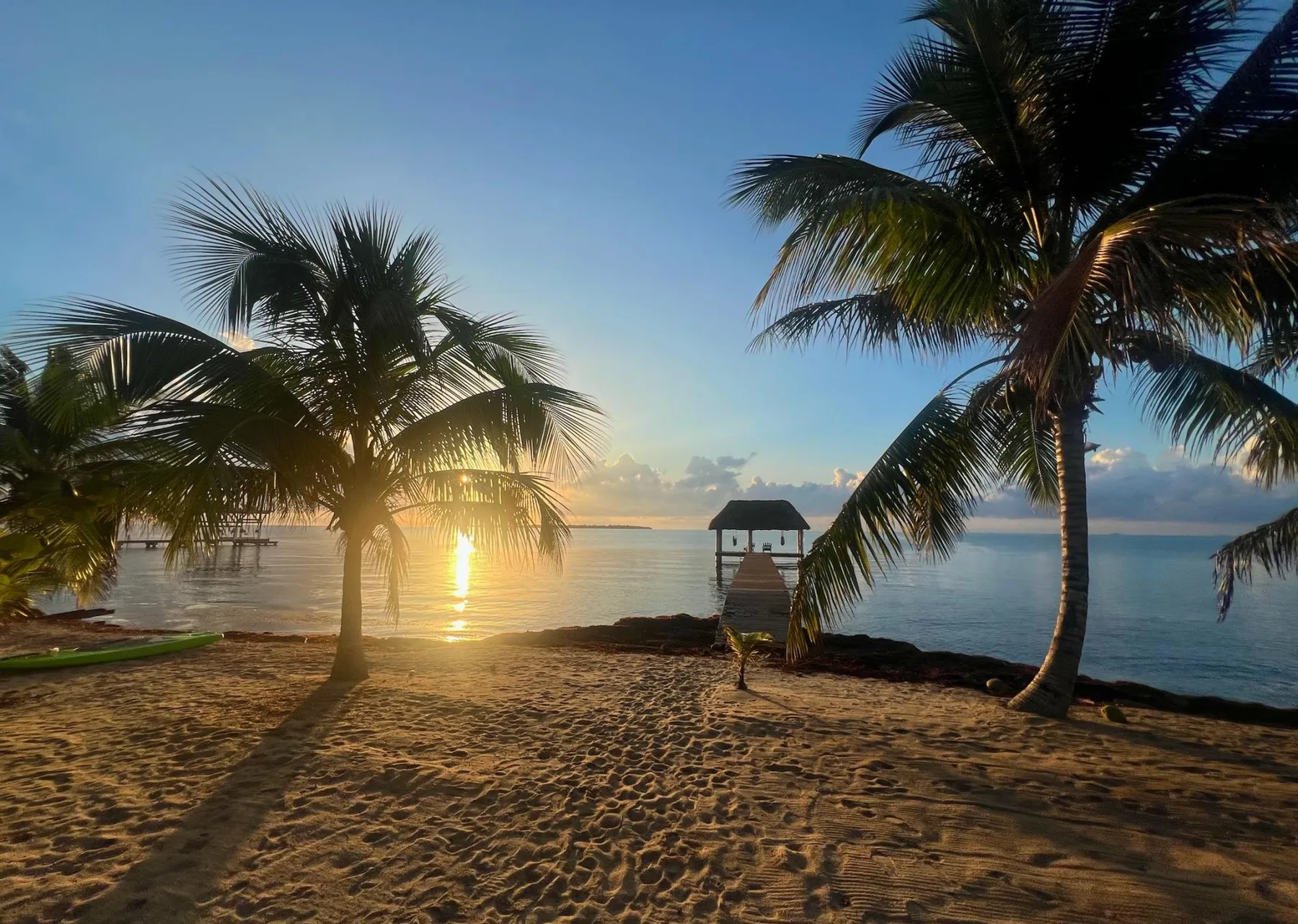 A sandy beach with palm trees frames a small pavilion over calm water at sunset.