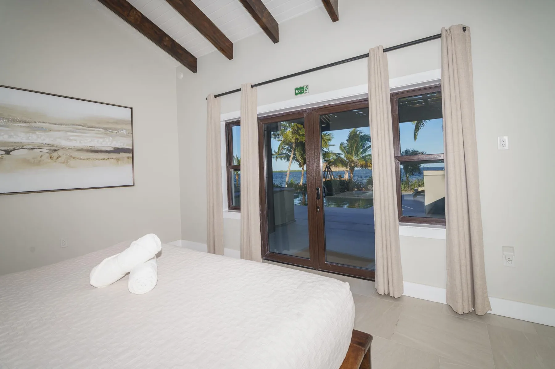 A bedroom with a bed beside glass doors overlooking palm trees and water.
