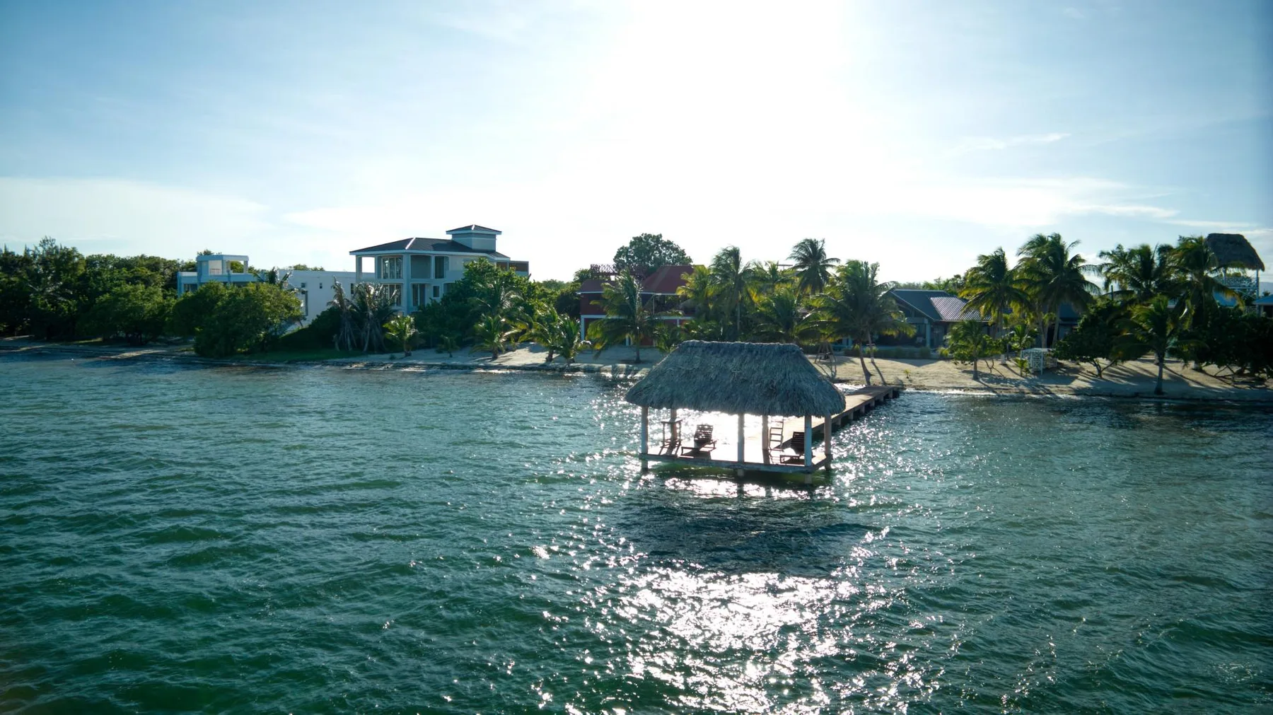 A thatched pavilion stands over the water near a tropical shoreline.