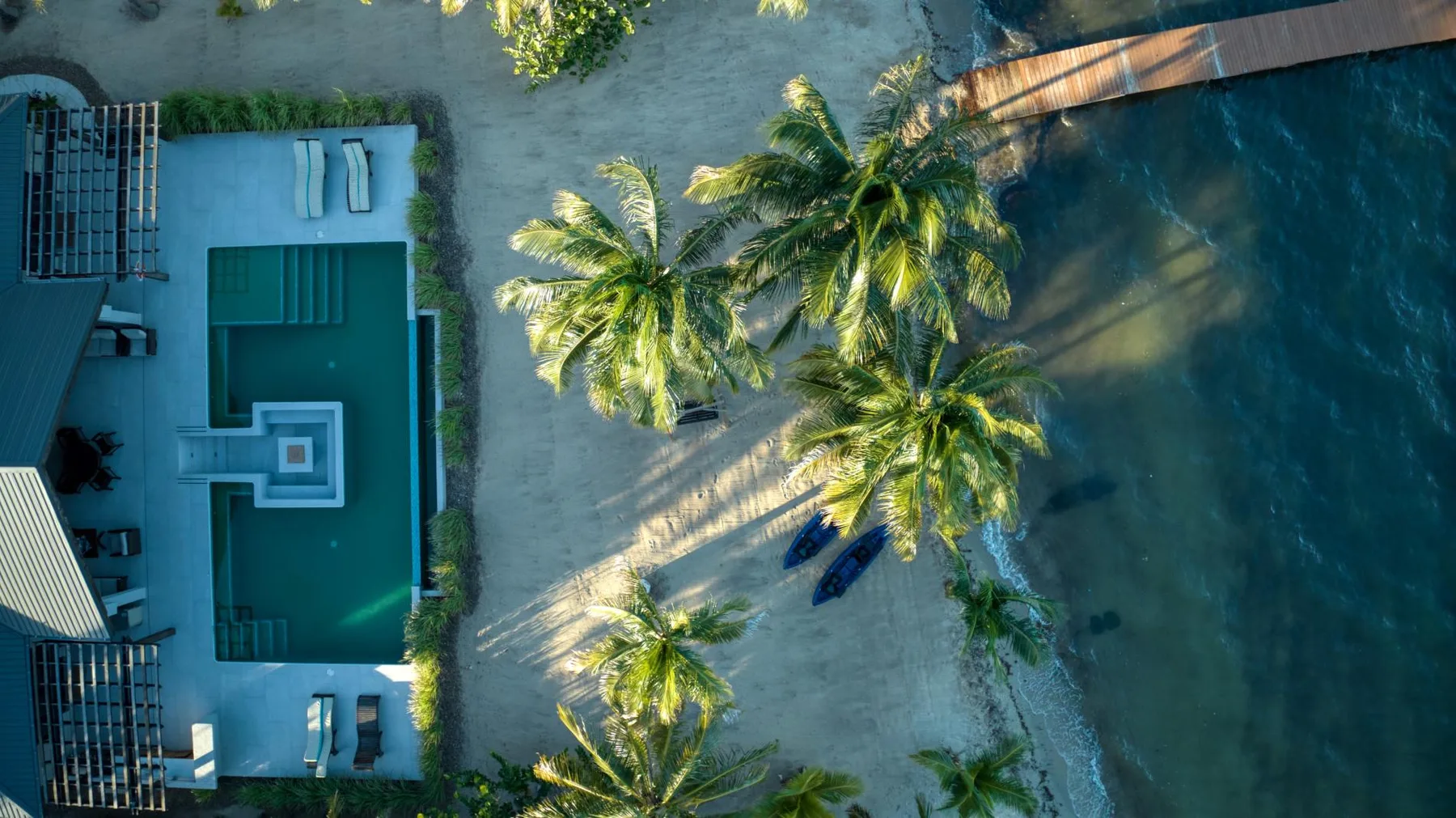 An aerial view shows a swimming pool beside a sandy beach and palm trees.