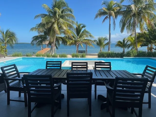 An outdoor dining area beside a swimming pool, with palm trees and water beyond.