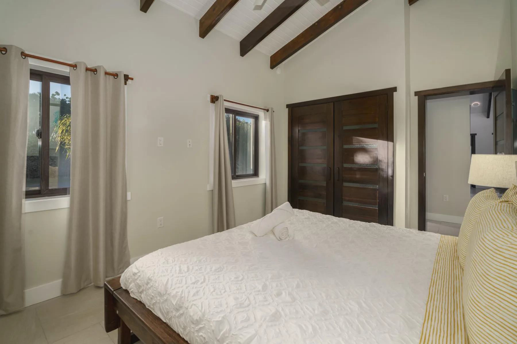 A bedroom with a bed, curtained windows, and exposed ceiling beams.