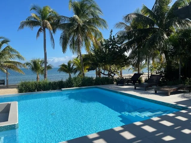 A swimming pool sits beside a palm-lined beach, with lounge seating nearby.